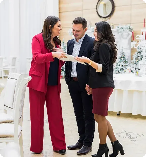 Three professionally dressed people, two women and one man, standing indoors in a decorated room, reviewing a document together and smiling.