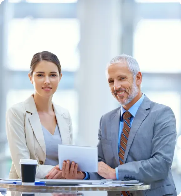 Professional man and woman in business attire sitting at a table with a tablet and coffee cup, smiling at the camera in a bright office.